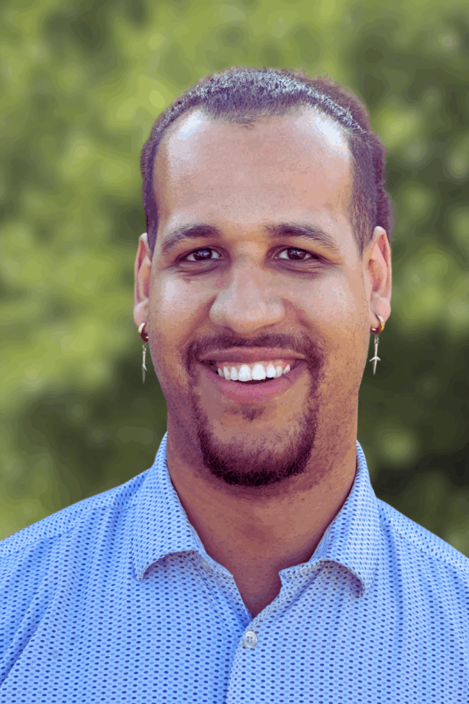 Smiling man with earrings in a blue shirt outdoors.