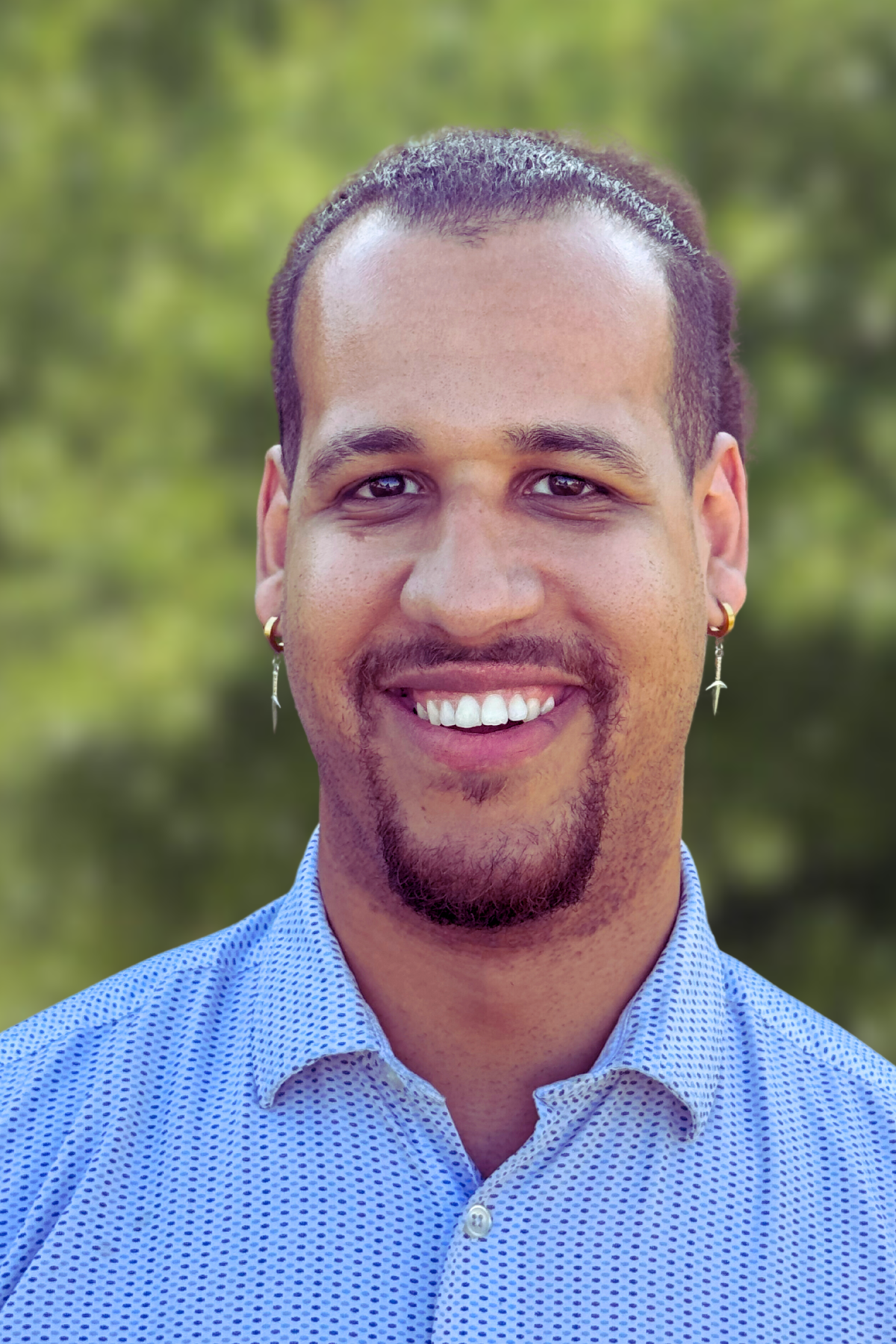 Smiling man with earrings in a blue shirt outdoors.